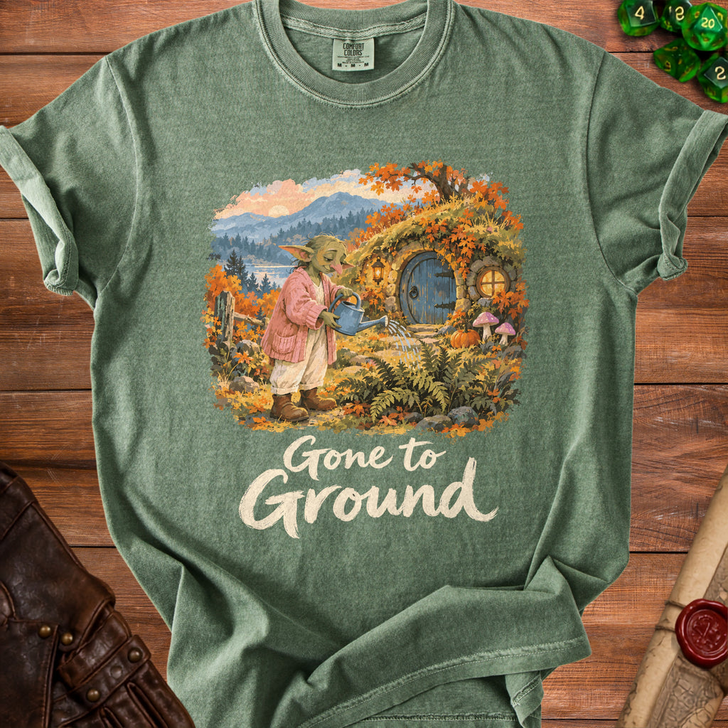 Green t-shirt featuring a creature watering a hobbit-like home with "Gone to Ground" text.