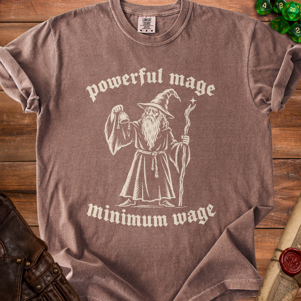 Brown t-shirt featuring a wizard illustration and the text powerful mage minimum wage, laid on wood.