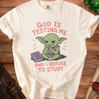 Goblin character on a cream t-shirt reads "God is testing me and I refuse to study."