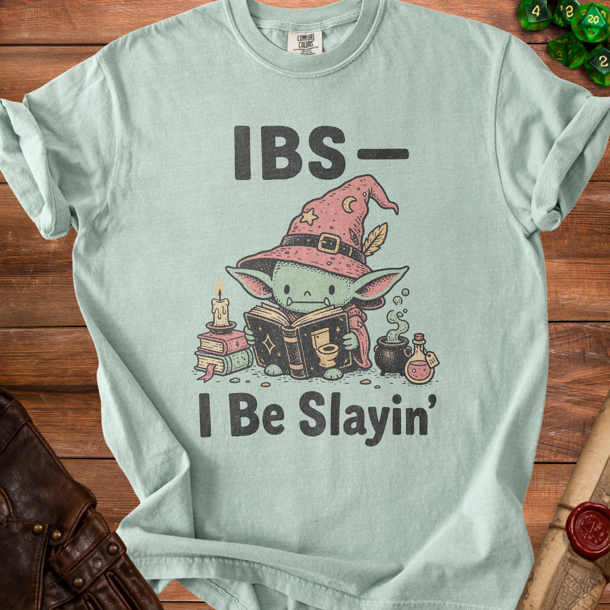 Light blue t-shirt with goblin character reading a book, wearing a witch hat, text reads "IBS- I Be Slain'".
