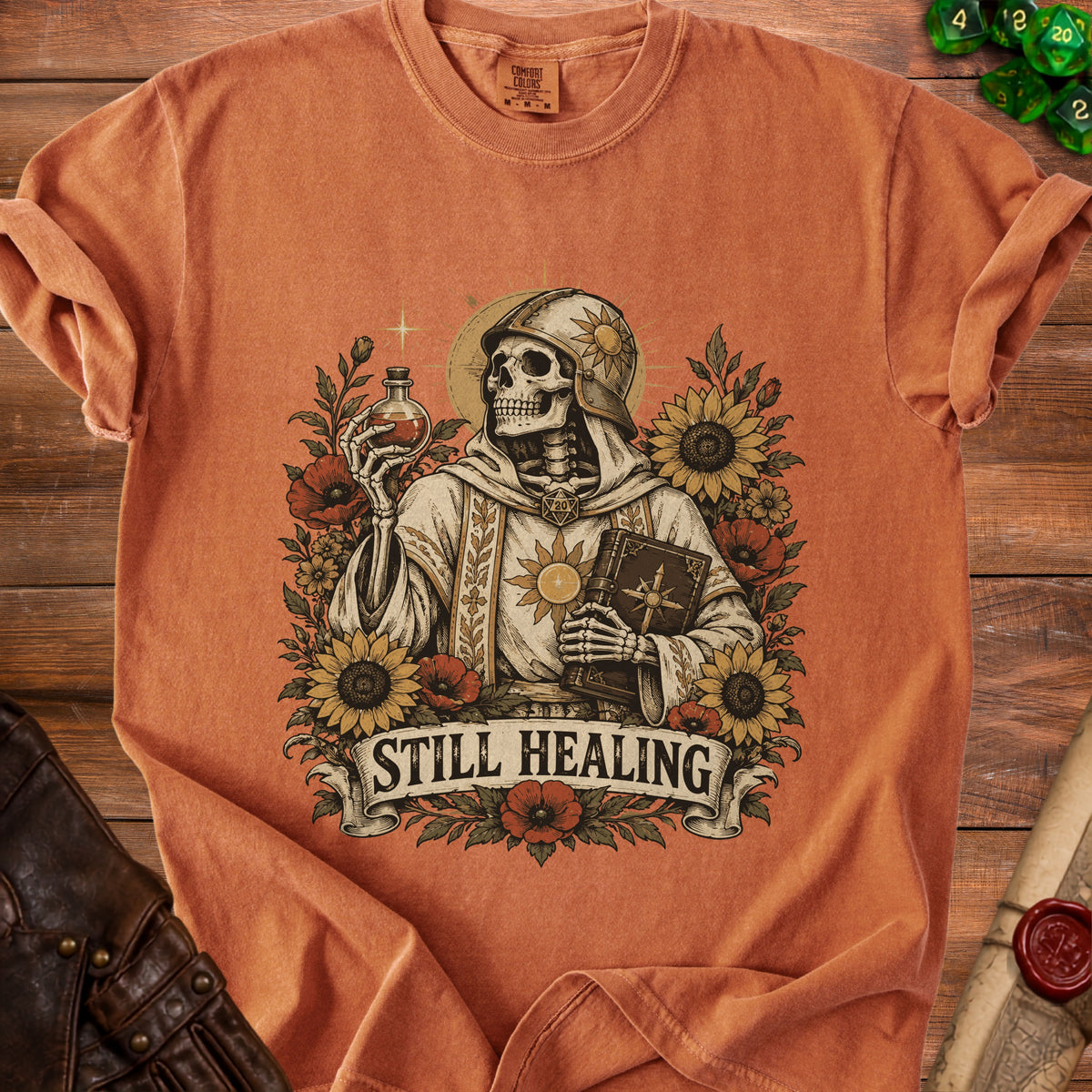 Orange t-shirt with skeleton holding potion and book surrounded by flowers and text "STILL HEALING".