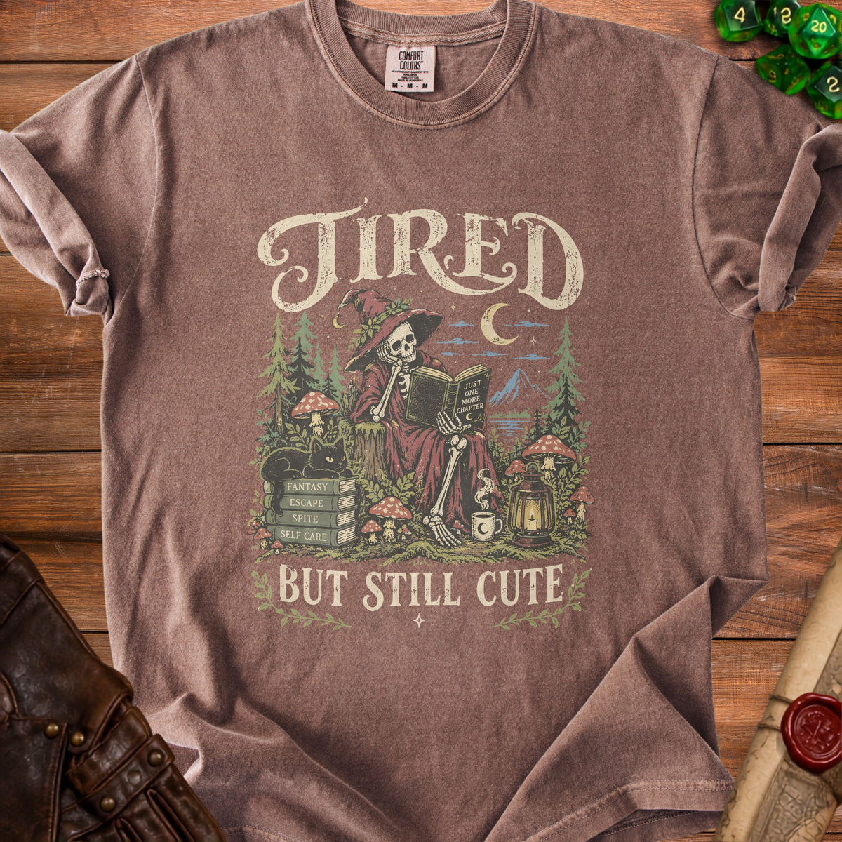Brown t-shirt with "Tired But Still Cute" skeleton reading a book graphic.