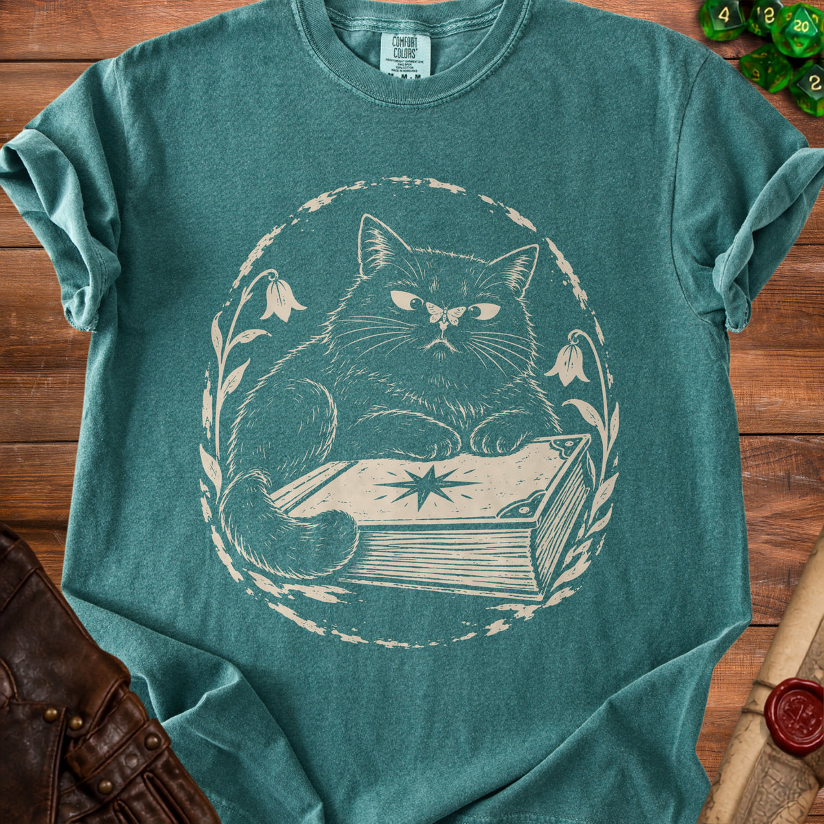 Teal t-shirt with a cream graphic of a cat on a book, surrounded by flowers.