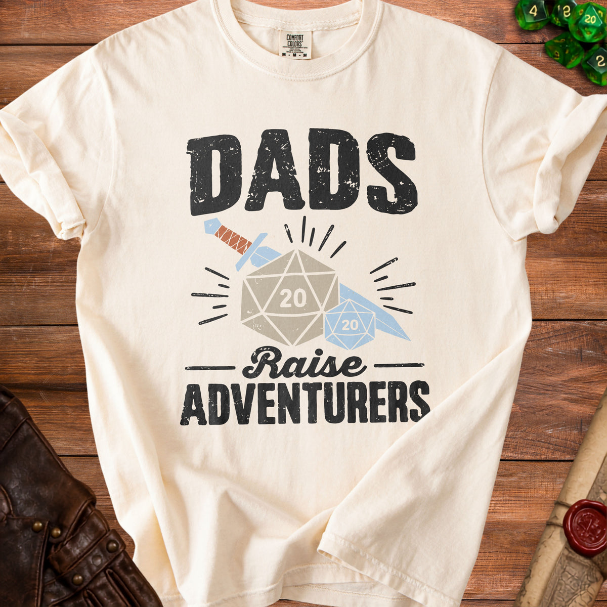 Cream t-shirt with "Dads Raise Adventurers" text, dice, and sword graphic.