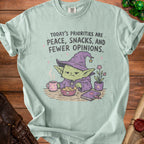 Mint green t-shirt with illustration of a grumpy goblin wearing a witch hat, reading, eating snacks, and drinking coffee.
