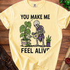 Yellow t-shirt with skeleton watering plants and text "YOU MAKE ME FEEL ALIVE."