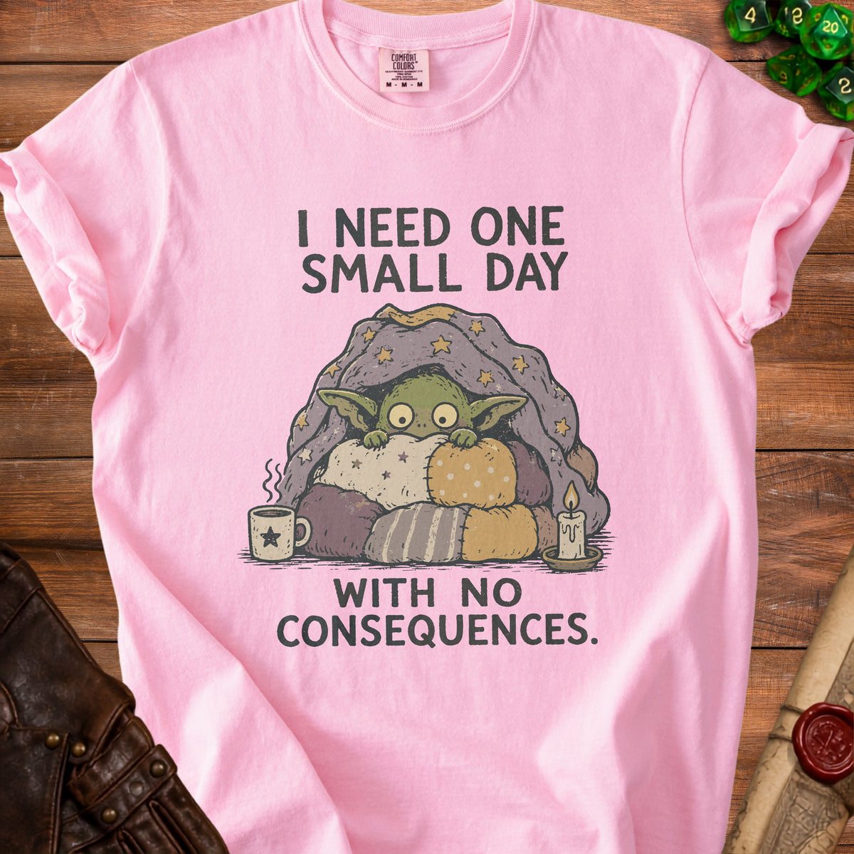 Pink t-shirt with a character wrapped in a blanket saying "I need one small day with no consequences."