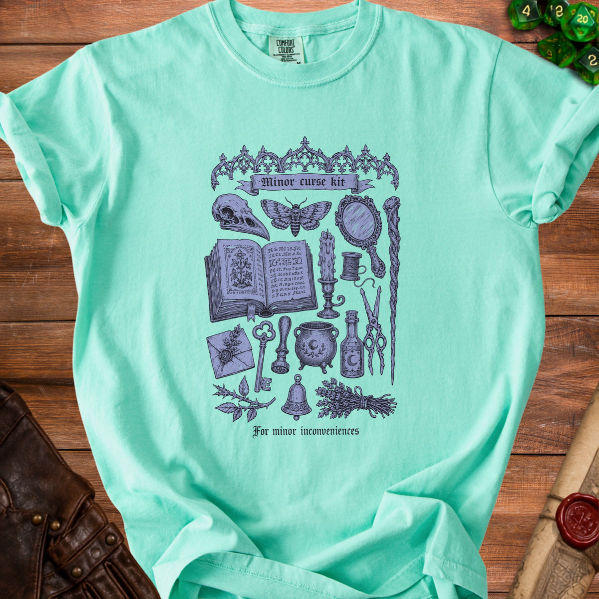 Light teal t-shirt with purple gothic illustration of spellbook, skull, moth, and cauldron titled "Minor curse kit".
