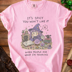 Pink t-shirt with illustration of a witch, black cat, spell books, and text "It's spicy you won't like it when people ask what I'm thinking".