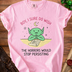 Pink t-shirt with a green cartoon creature and the text "Boy, I sure do wish the horrors would stop persisting."