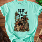 Skeleton in witch hat reading with black cat, "Rest is a Spell" text.