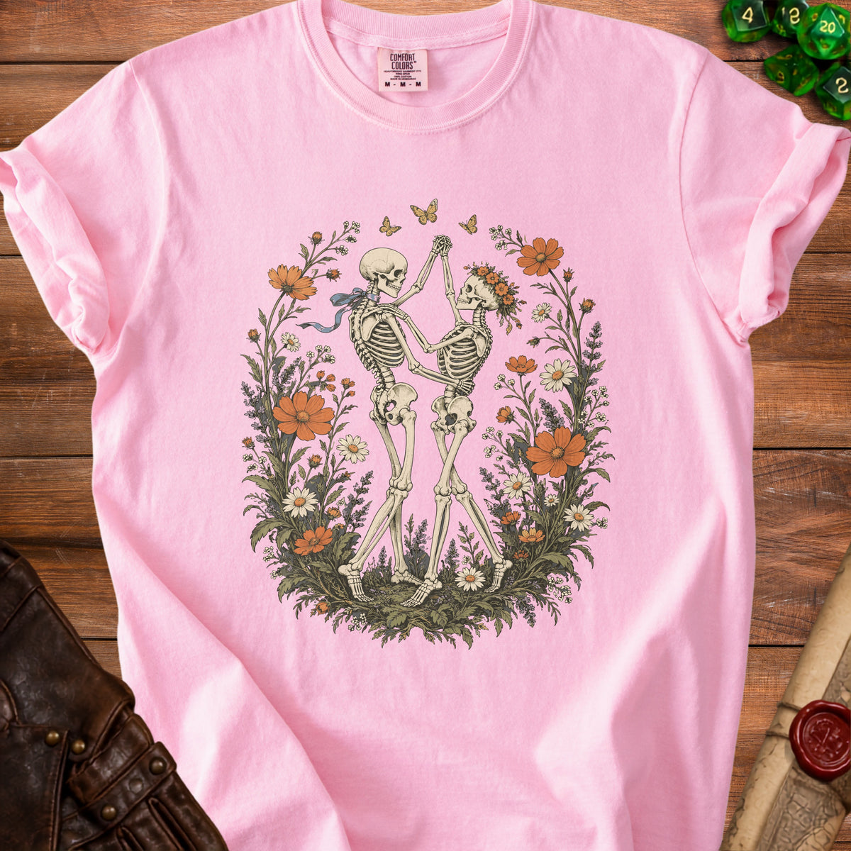 A pink t-shirt featuring a graphic of two dancing skeletons surrounded by flowers and butterflies.