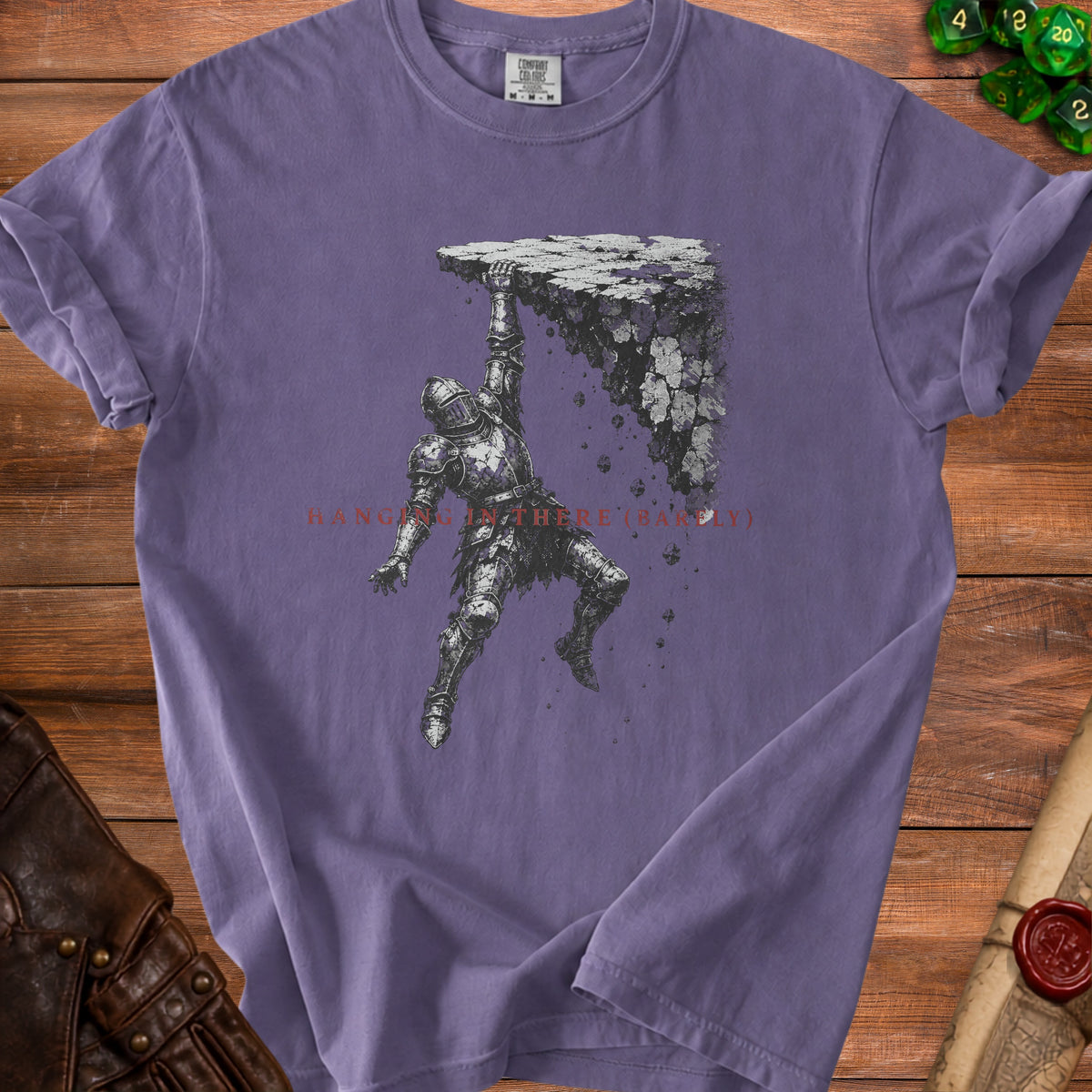 Purple t-shirt with a knight hanging from crumbling rocks and text "hanging in there (barely)".