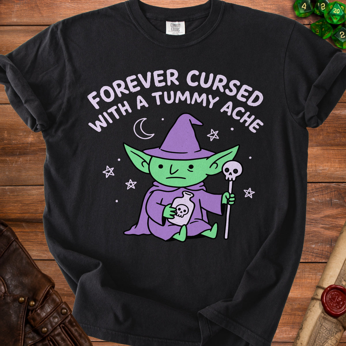 Black t-shirt with a green creature holding a potion bottle and text "FOREVER CURSED WITH A TUMMY ACHE".