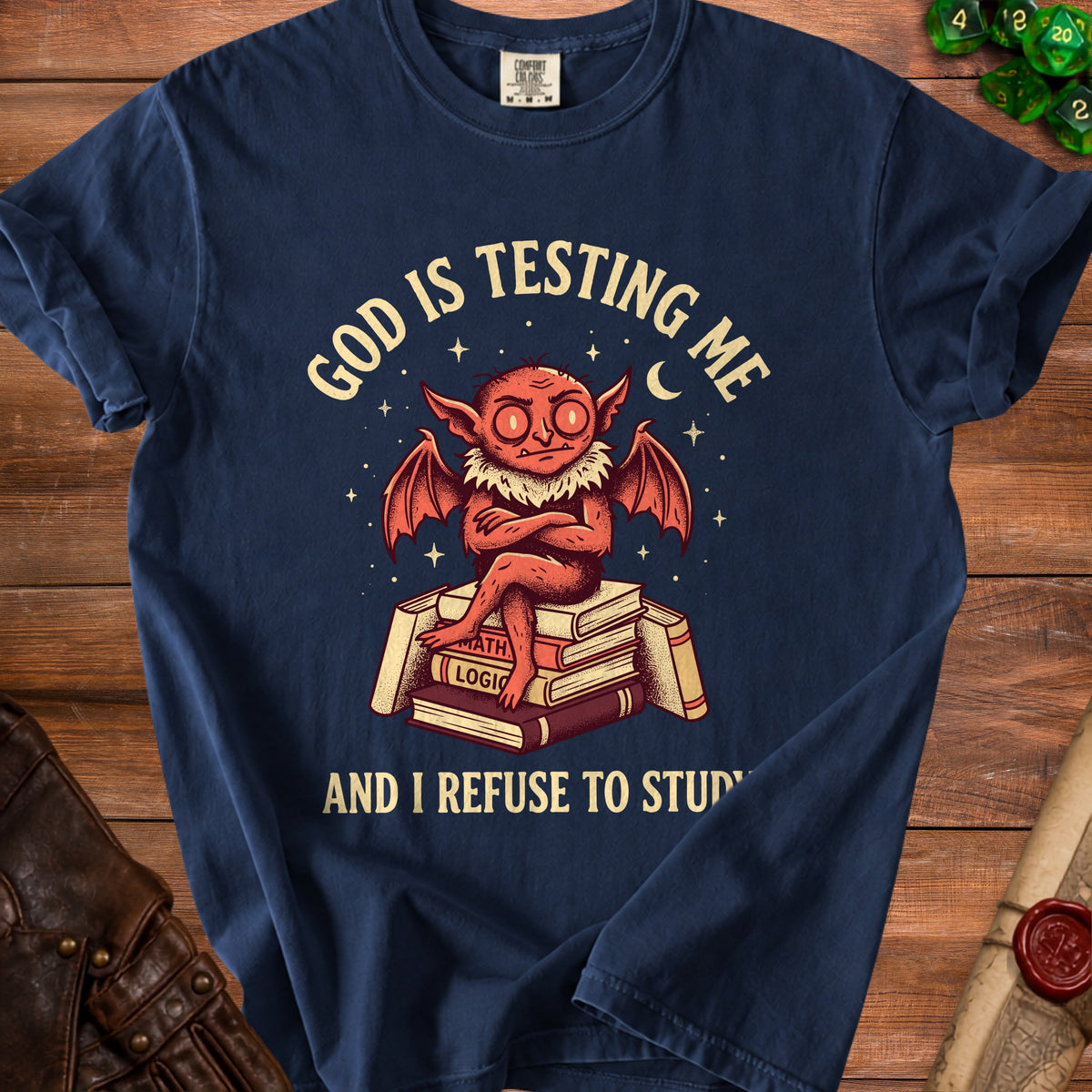 Navy t-shirt with a red winged creature sitting on books, reading "God is testing me and I refuse to study."