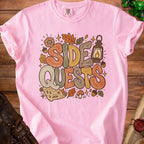 Pink t-shirt with "Side Quests" graphic featuring dice, lantern, map, and fall leaves.