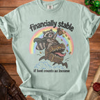 Light teal t-shirt with raccoon on treasure chest, rainbow, and text "financially stable if loot counts as income".