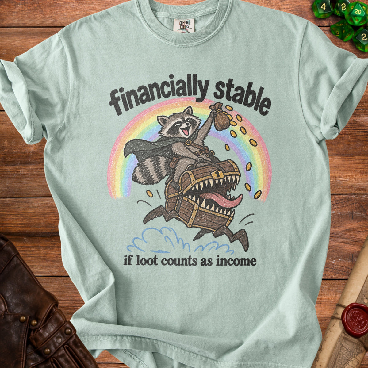Light teal t-shirt with raccoon on treasure chest, rainbow, and text "financially stable if loot counts as income".