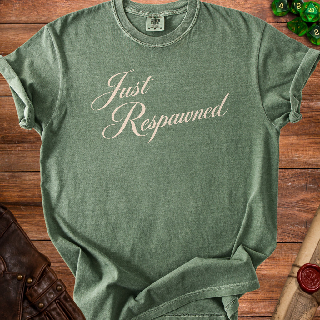 Green t-shirt with "Just Respawned" text, laid on a wooden table with dice and leather accents.