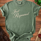 Green t-shirt with "Just Respawned" text, laid on a wooden table with dice and leather accents.
