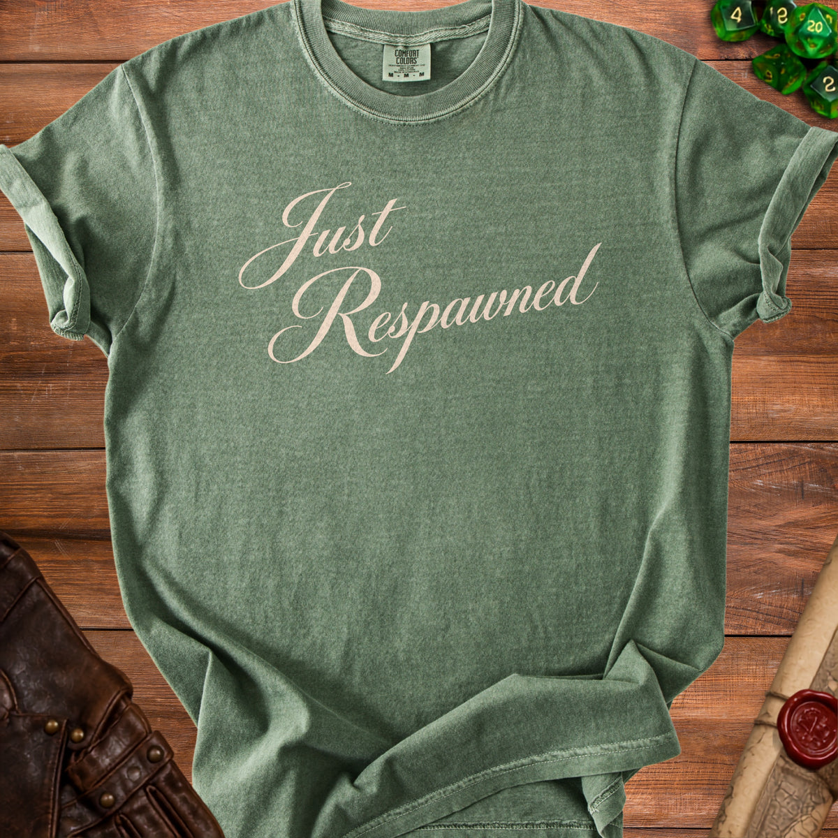 Green t-shirt with "Just Respawned" text, laid on a wooden table with dice and leather accents.