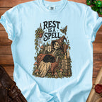 Skeleton relaxing with black cat, text "REST IS A SPELL," "STILL HERE," "LESS DOING MORE BEING."