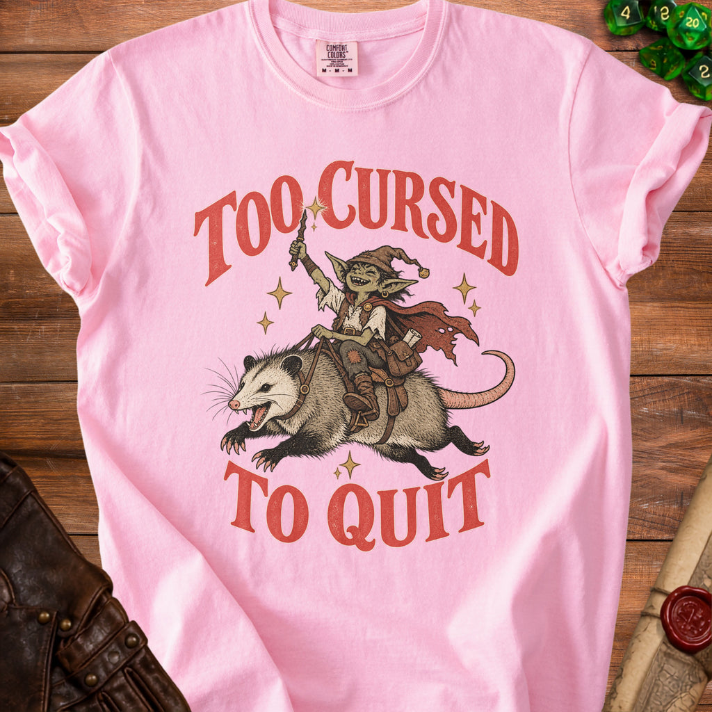 Pink t-shirt with a goblin riding a possum and text "Too Cursed To Quit".