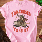 Pink t-shirt with a goblin riding a possum and text "Too Cursed To Quit".