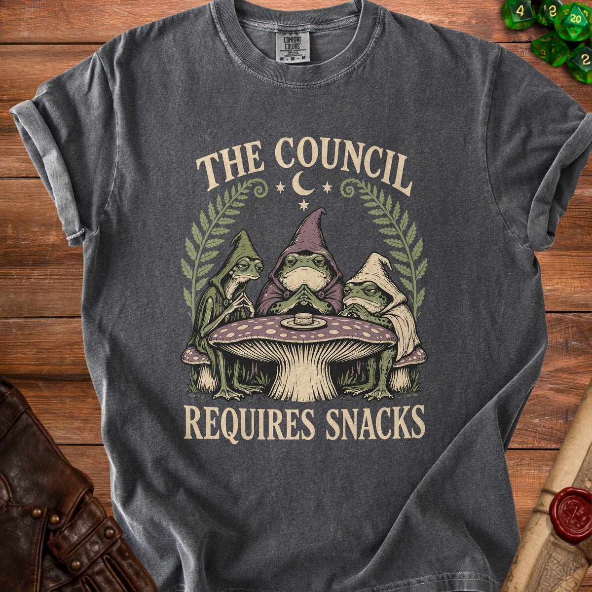 Dark grey t-shirt with a graphic of three hooded frogs at a mushroom table, text "The Council Requires Snacks".
