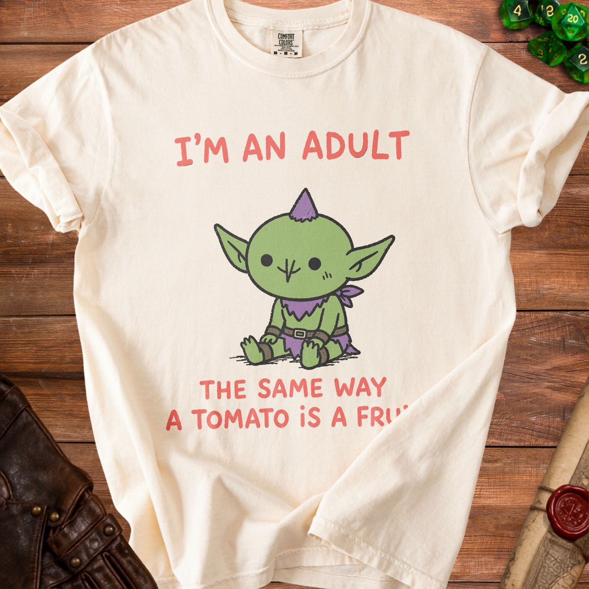 Cream t-shirt with cartoon goblin and text "I'm an adult the same way a tomato is a fruit."