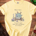 Yellow shirt with illustration of a creature writing and text "Back then I didn't even have the confidence to dream."