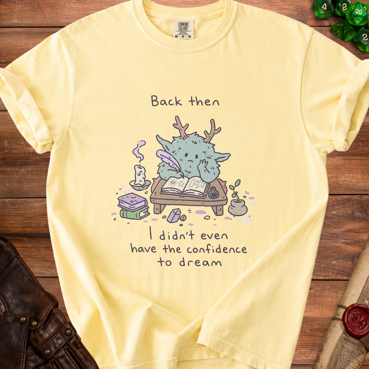 Yellow shirt with illustration of a creature writing and text "Back then I didn't even have the confidence to dream."