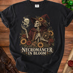 A black t-shirt features a graphic of a skeleton wearing a witch hat, surrounded by flowers, with the text NECROMANCER IN BLOOM.