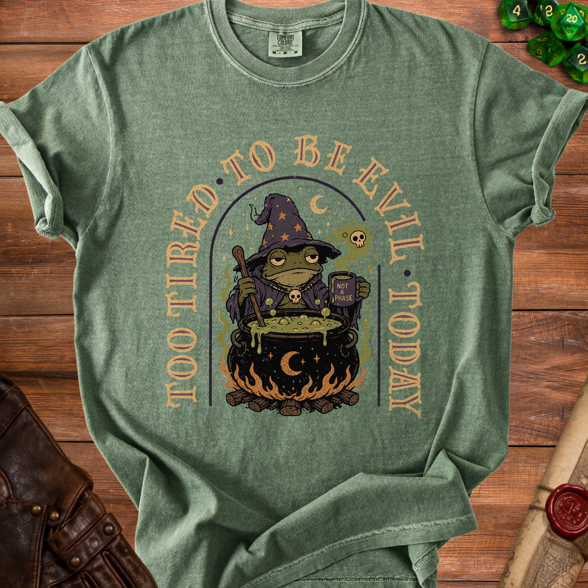 Green t-shirt graphic with a frog wizard and cauldron, reading "Too Tired To Be Evil Today".