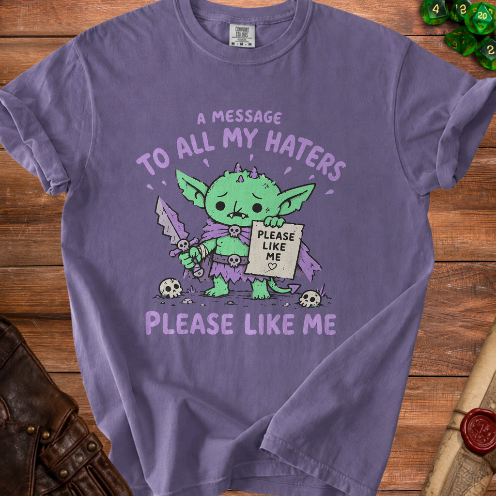 Purple t-shirt with a sad green goblin holding a sword and "Please Like Me" sign.