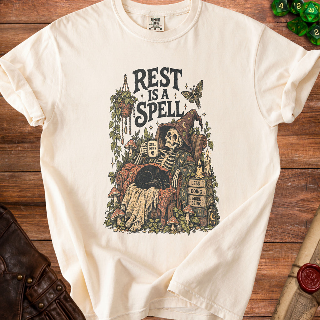 Cream t-shirt graphic of skeleton resting with a cat, "Rest is a Spell" text.
