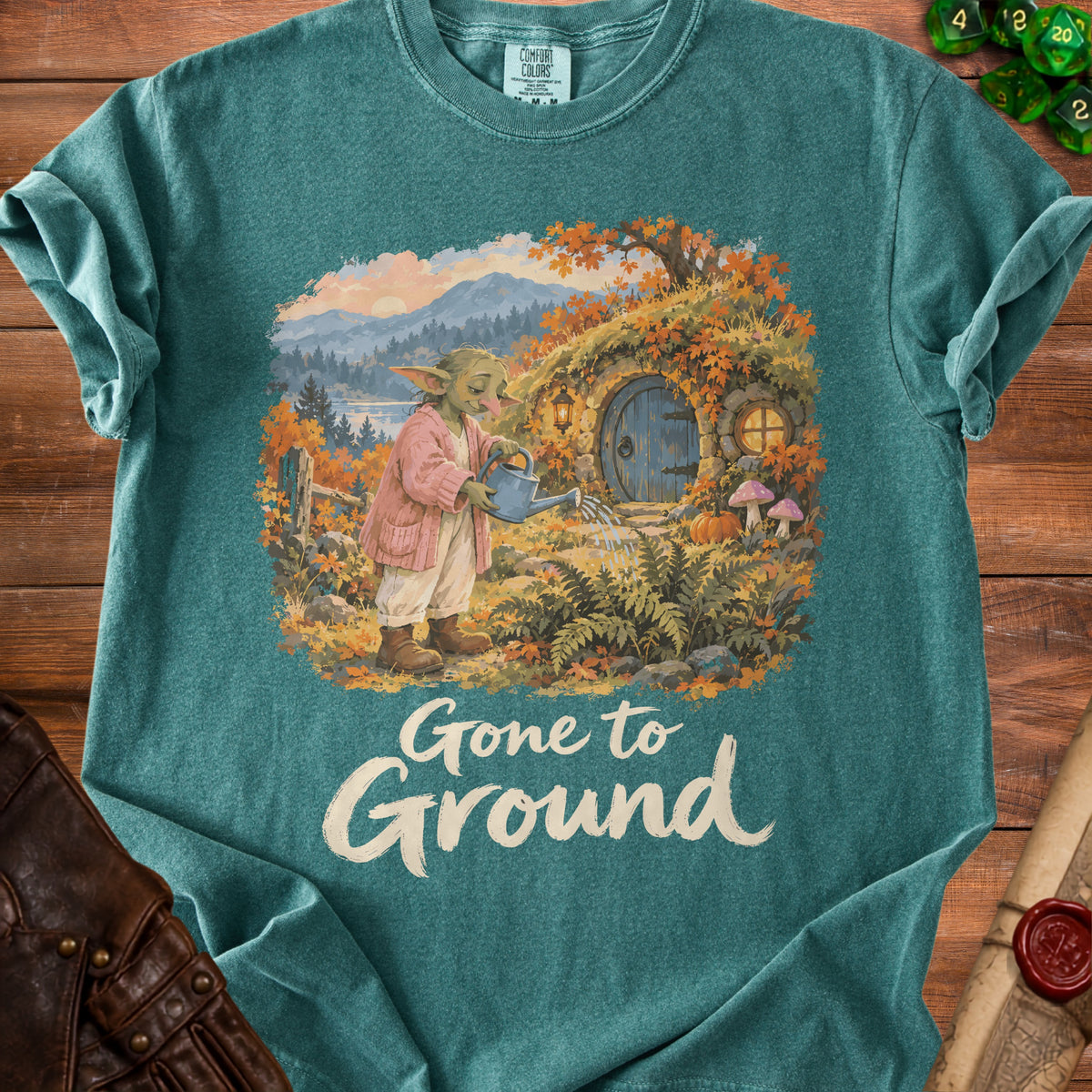 T-shirt graphic of a green creature watering plants outside a hobbit-like home.
