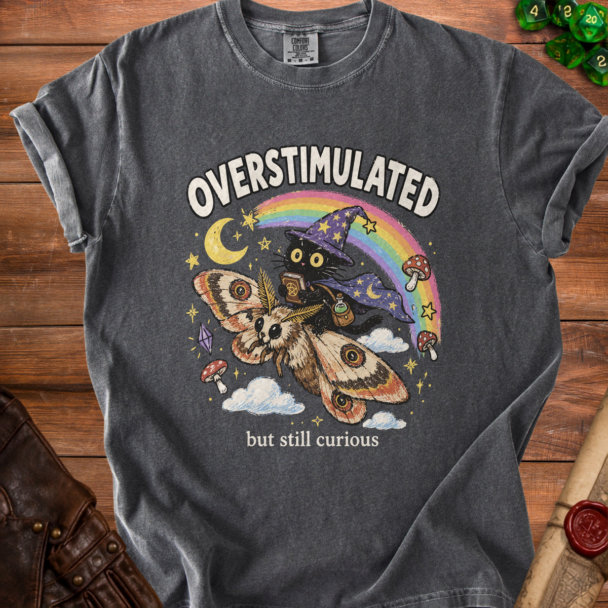 Dark t-shirt with a graphic of a black cat riding a moth, with "OVERSTIMULATED but still curious".