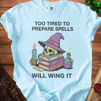 Light blue t-shirt with a skull wearing a wizard hat, books, potion, and candle graphic. Text: "TOO TIRED TO PREPARE SPELLS WILL WING IT".