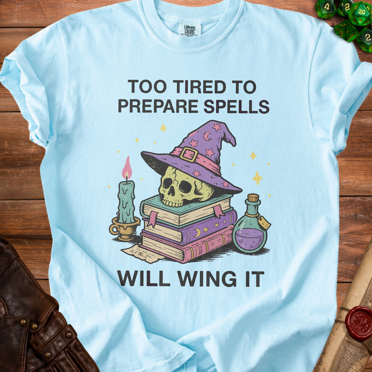 Light blue t-shirt with a skull wearing a wizard hat, books, potion, and candle graphic. Text: "TOO TIRED TO PREPARE SPELLS WILL WING IT".