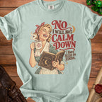 Light blue t-shirt with graphic of woman holding coffee and a book, "No I will not calm down, I have hexes to finish."