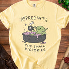 Yellow t-shirt with Yoda-like character in bathtub holding skull mug. Text reads "APPRECIATE THE SMALL VICTORIES".