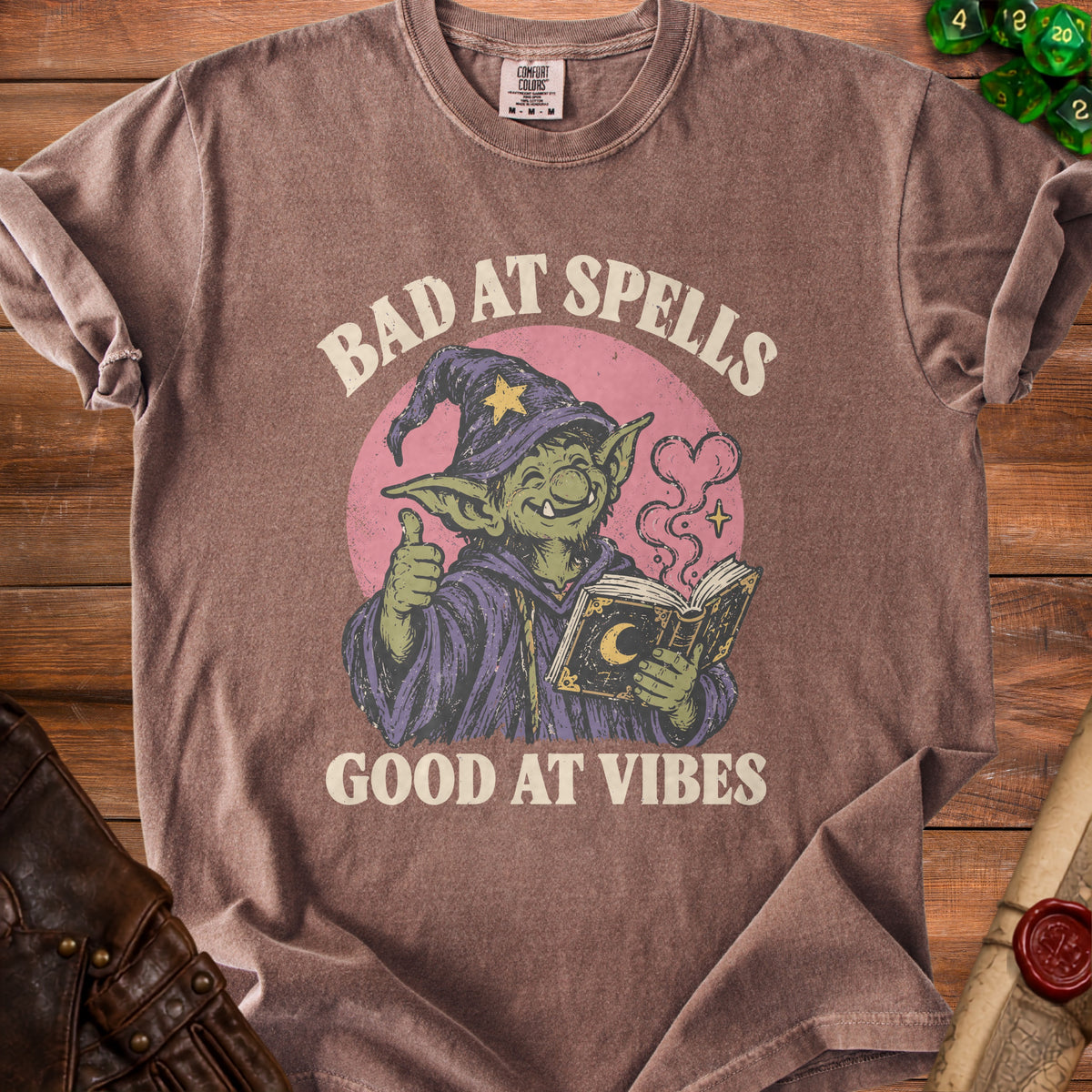 Brown t-shirt with a green goblin wizard graphic and text "BAD AT SPELLS GOOD AT VIBES".