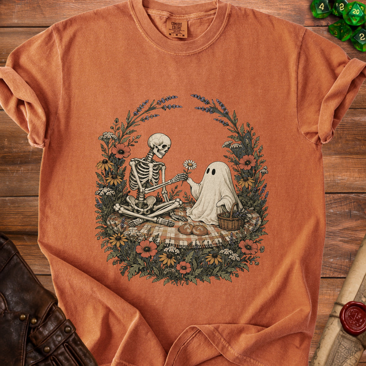 Orange t-shirt featuring an illustration of a skeleton and a ghost having a picnic surrounded by flowers.