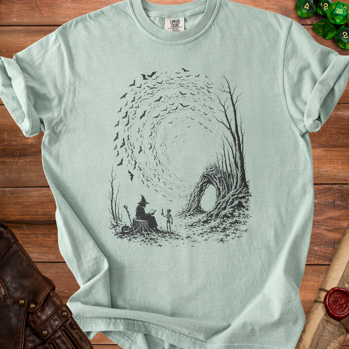 Light green t-shirt with black illustration of a witch, skeleton, and bats.