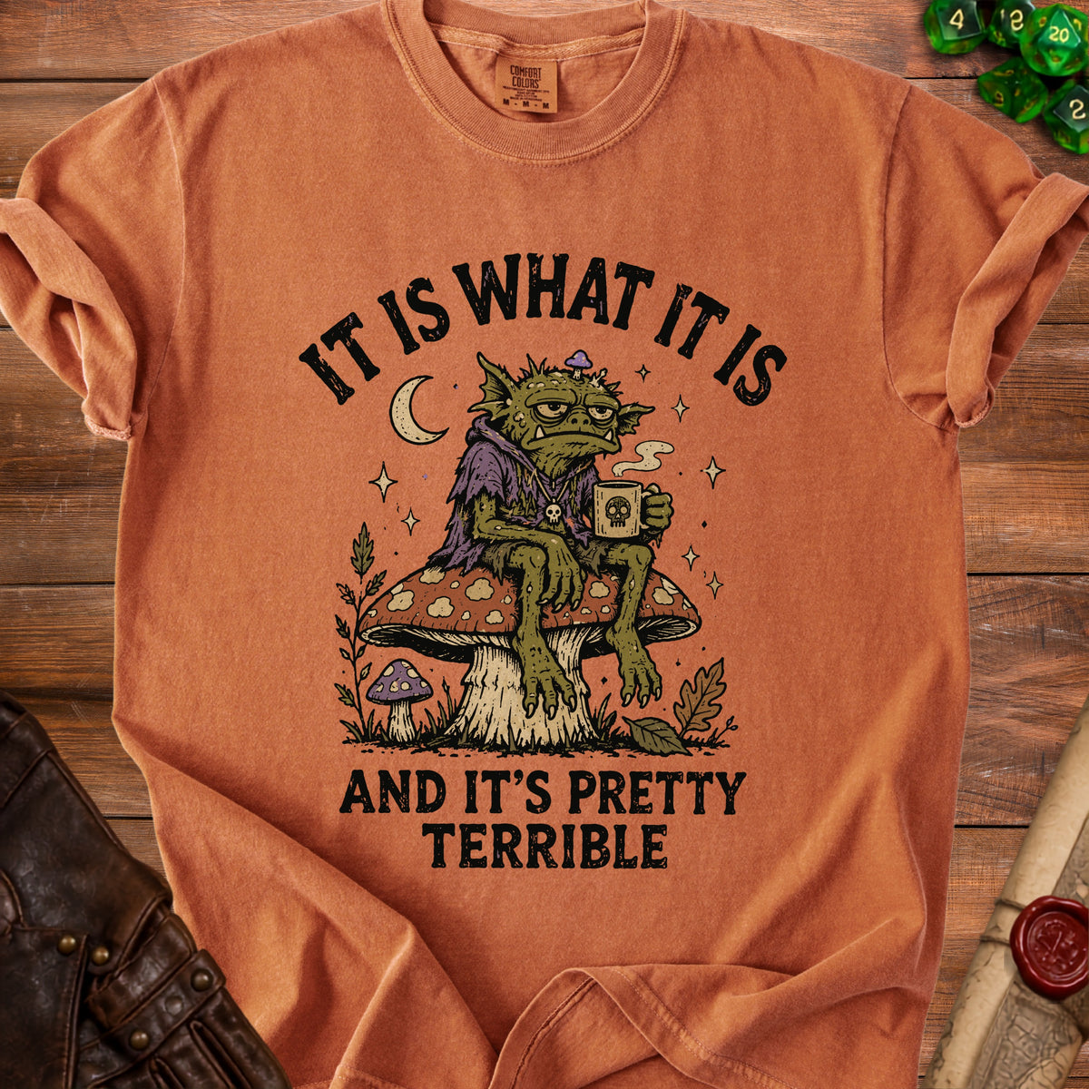 Orange t-shirt with graphic of a monster sitting on a mushroom, drinking coffee. Text reads "It is what it is and it's pretty terrible."