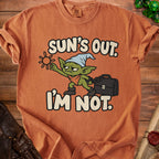 Orange t-shirt with cartoon goblin holding sun and briefcase, "Sun's Out. I'm Not." text.