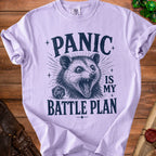 Light purple t-shirt with a distressed print of a possum, a D20 die, and the text "PANIC IS MY BATTLE PLAN".