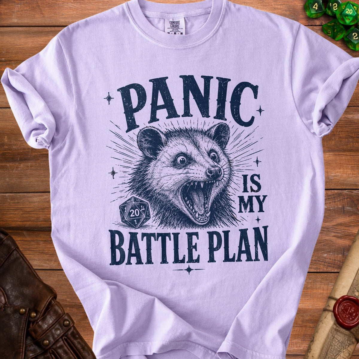 Light purple t-shirt with a distressed print of a possum, a D20 die, and the text "PANIC IS MY BATTLE PLAN".