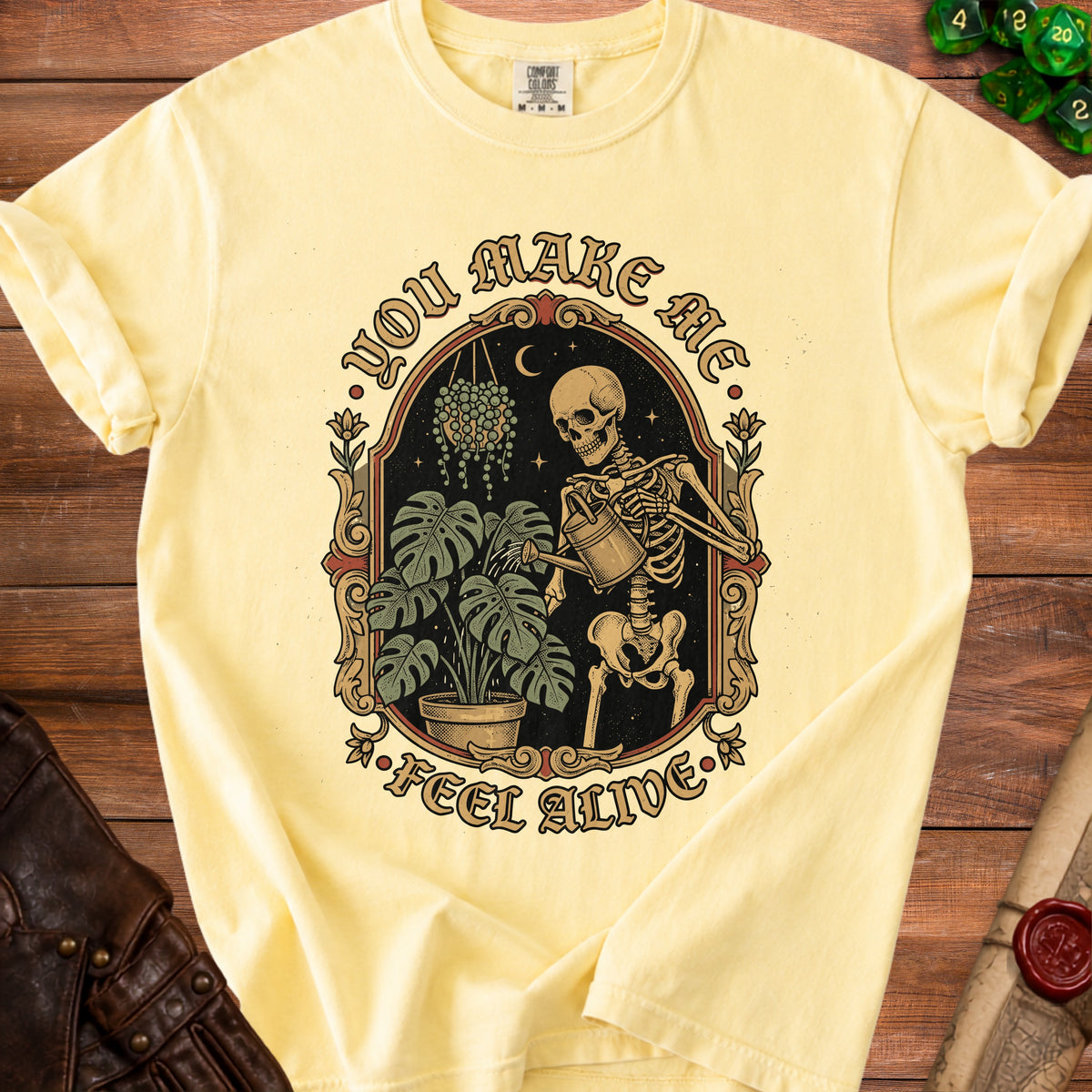 Light yellow t-shirt with skeleton watering plants and text "You Make Me Feel Alive."